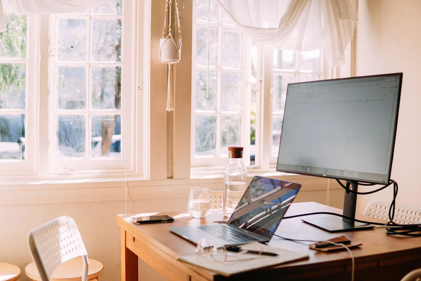 Laptop, monitor and glasses on a wooden desk beside a sunlit window.