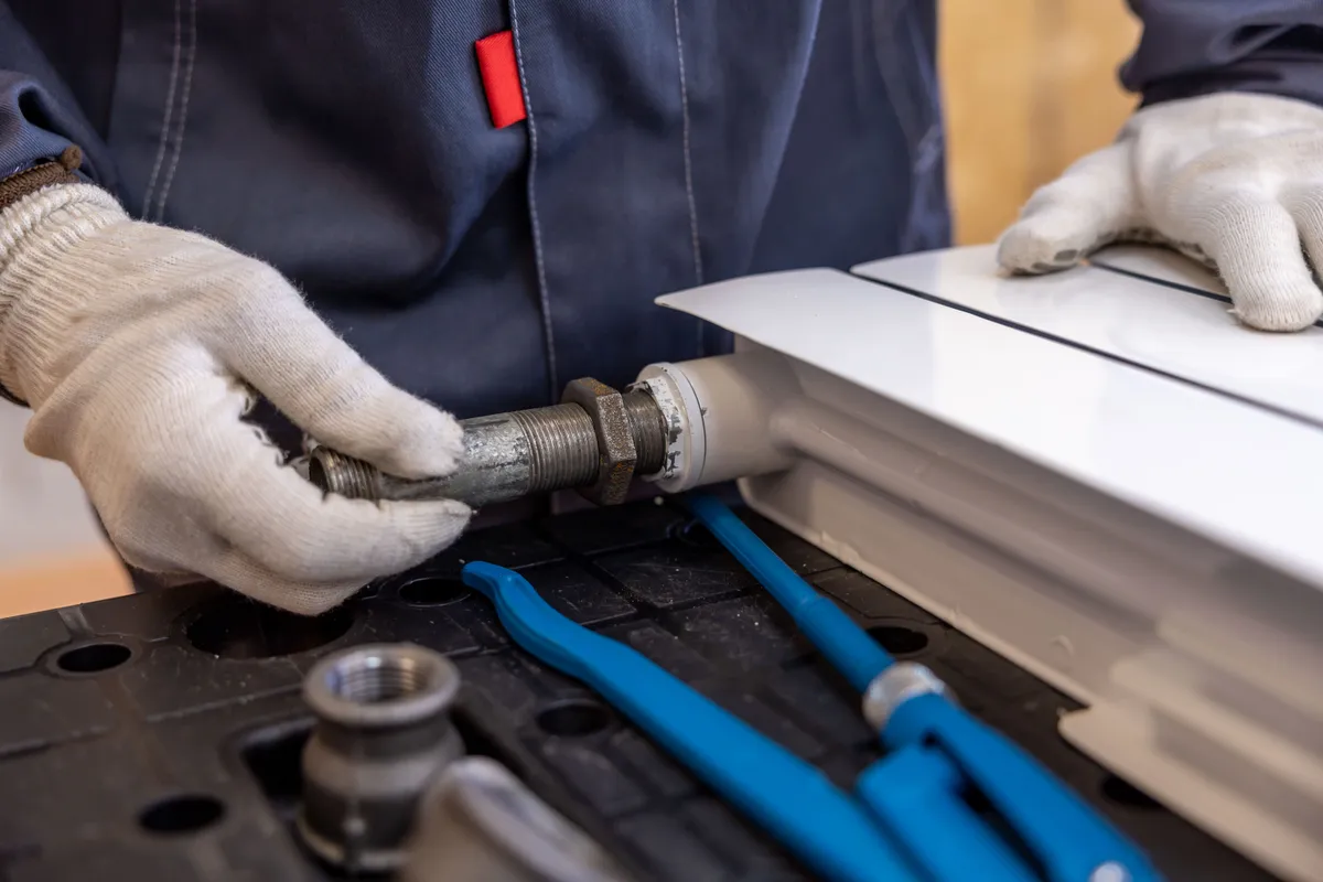 A plumber's gloved hands fitting a radiator pipe with hand tools